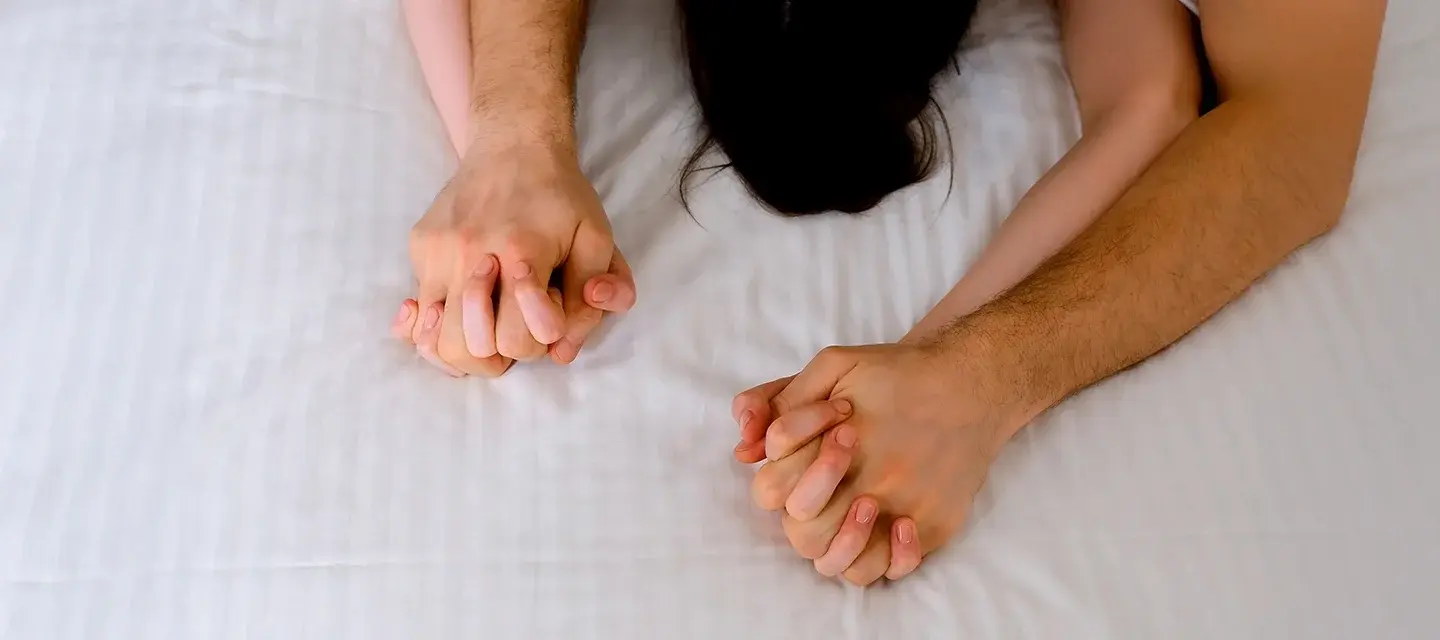 A couple clutching hands during sex on white sheets